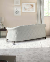 Elegant contemporary off-white sofa with a tufted, diamond-patterned backrest and sloping arms, seated on dark wooden legs, stationed on a textured off-white area rug.