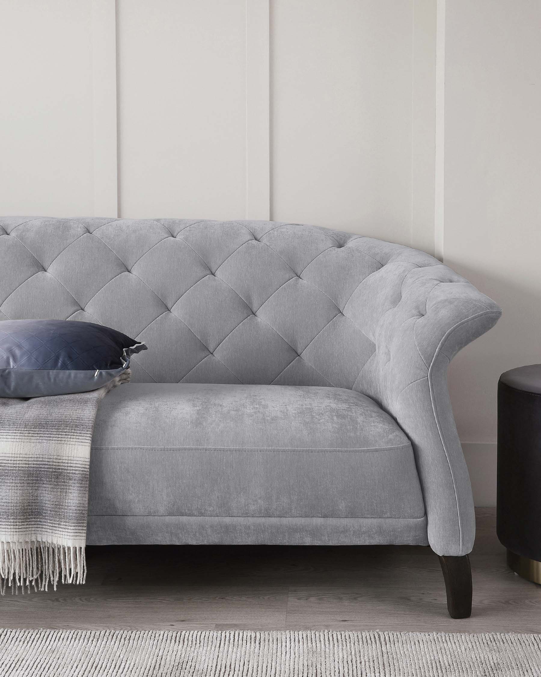 Elegant grey tufted chaise lounge with curved backrest, sophisticated diamond stitching, velvety upholstery, and dark wooden legs, complemented by a dark blue throw pillow and a light grey throw blanket with fringe detailing.