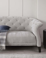 Elegant light-grey, fabric upholstered loveseat with tufted button detailing on the high backrest. The sofa features a curved back, smooth seat cushion, and dark-stained, tapered wooden legs. Accented with a navy blue decorative pillow and a light-grey throw with fringe detailing on the left side, placed upon a textured off-white rug.