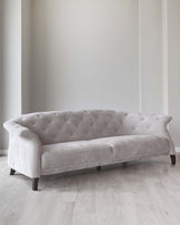 Elegant light grey upholstered sofa with tufted backrest and plush seating, featuring a curvaceous profile and dark wooden legs, positioned against a neutral wall on a light wooden floor.