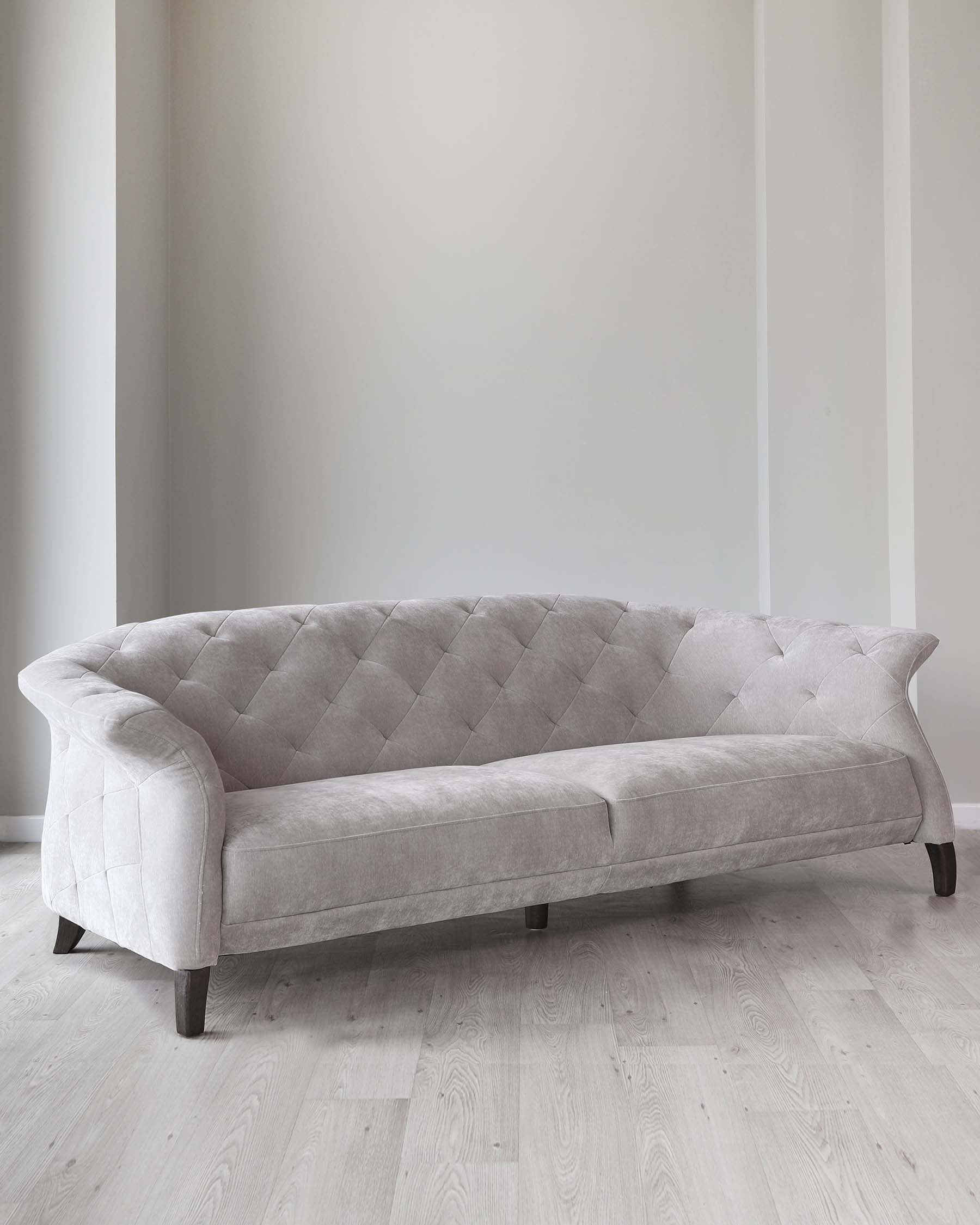 Elegant light grey upholstered sofa with tufted backrest and plush seating, featuring a curvaceous profile and dark wooden legs, positioned against a neutral wall on a light wooden floor.