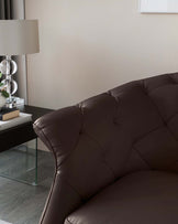 A luxurious chocolate brown leather sofa with a tufted backrest and smooth seat cushions, accompanied by a sleek glass-top side table with a metal frame, featuring a modern lamp with a translucent base and cylindrical shade, and a framed abstract wall art above.