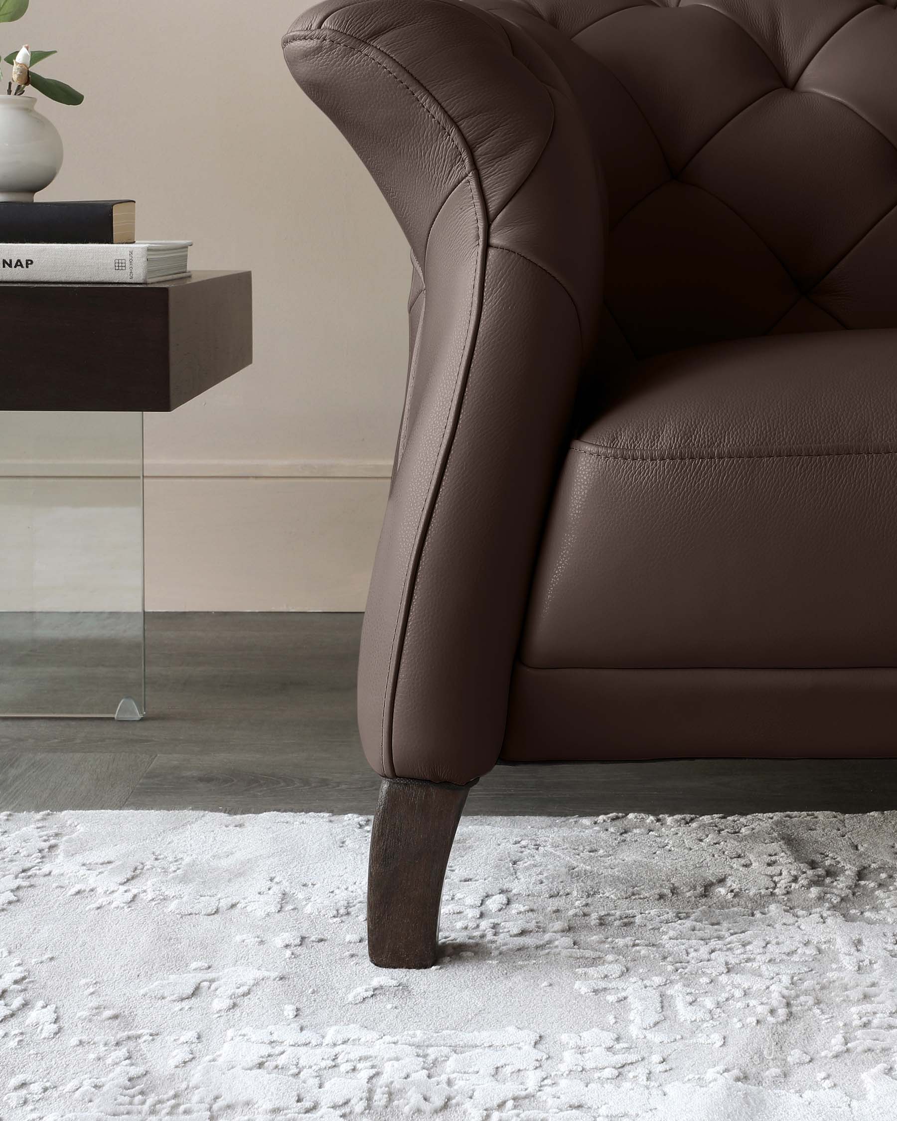 Close-up view of a luxurious brown leather armchair with tufted backrest, showcasing dark wooden legs, placed on a textured white rug. A modern side table with a dark finish and a minimalistic vase is partially visible in the background.