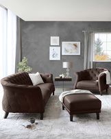 Elegant living room furniture set including a chocolate brown tufted leather sofa, matching armchair, and ottoman. A contemporary style side table with a lamp and decorative items. All pieces rest on a neutral-toned area rug with a distressed pattern.