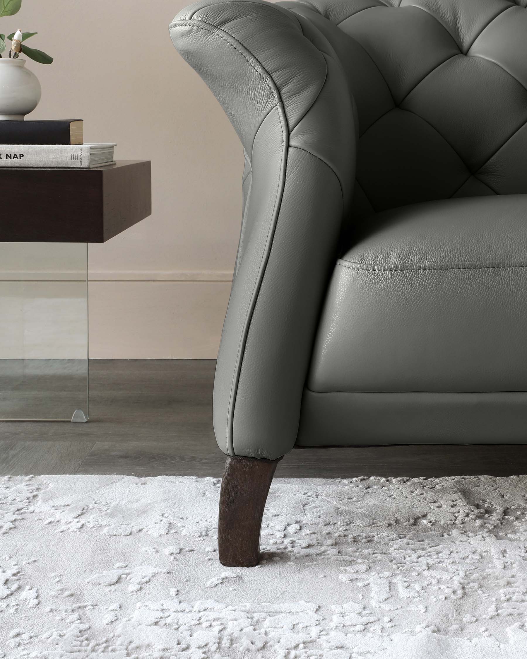 Elegant grey tufted leather armchair with a rolled arm design and dark wooden legs, complemented by a minimalist dark wooden side table holding books and a decorative plant, all above a textured white area rug.