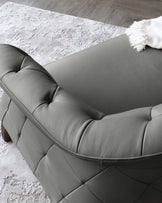 Modern grey tufted leather sofa with cylindrical wooden legs and a creamy white fringe throw blanket on an abstract-patterned white and grey area rug, set against a wood-finished flooring.