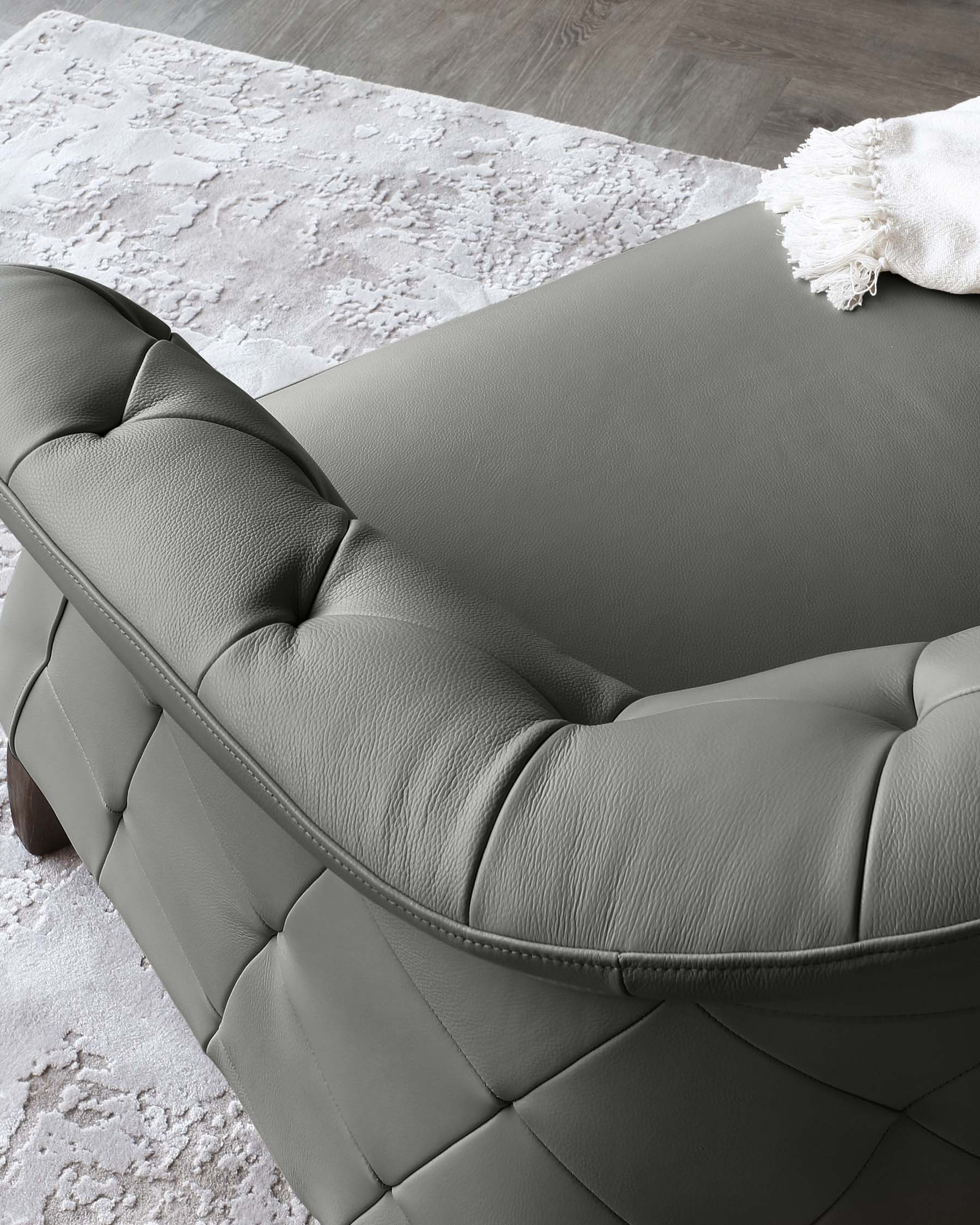 Elegant dark grey tufted leather sofa with a sophisticated silhouette, featuring plush cushioning, visible seam detailing, and wooden legs, partially draped with a cream fringed throw blanket, against a backdrop of a soft-textured white and grey area rug on a wooden floor.