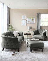 Elegant living room set including a tufted grey leather sofa with a matching armchair and ottoman. The pieces feature a diamond-stitched pattern and are complemented by dark wooden legs. A rectangular wooden side table with a bottom shelf and a square white tabletop lamp are also present.