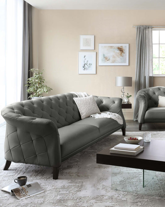 Elegant living room set featuring a tufted dark grey sofa with matching armchair, complemented by a modern dark brown rectangular coffee table with a glass bottom shelf. The room is accented with a soft textured area rug, simplistic white-and-silver decorative pillows, and a small round side table supporting a contemporary lamp with a grey shade.