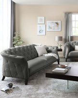 Elegant living room featuring a tufted dark grey upholstered sofa with curved arms and matching armchair, complemented by a sleek rectangular dark wood coffee table with a transparent glass base. The room is accented with a textured off-white area rug and a modern table lamp with a grey shade on a small side table. Decorative elements include various framed wall art, a white knitted throw pillow, and a ceramic vase with greenery.