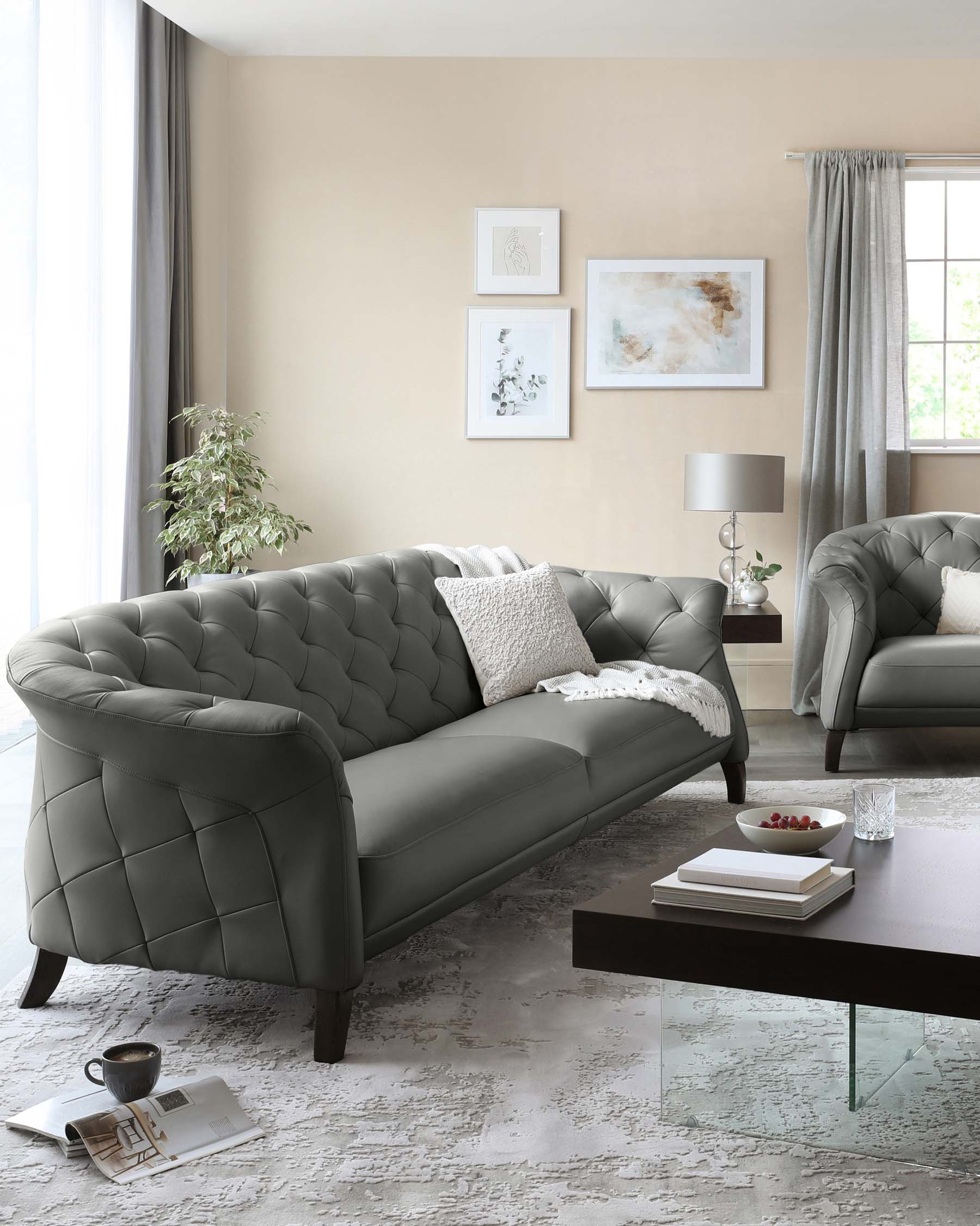 Elegant living room featuring a tufted dark grey upholstered sofa with curved arms and matching armchair, complemented by a sleek rectangular dark wood coffee table with a transparent glass base. The room is accented with a textured off-white area rug and a modern table lamp with a grey shade on a small side table. Decorative elements include various framed wall art, a white knitted throw pillow, and a ceramic vase with greenery.