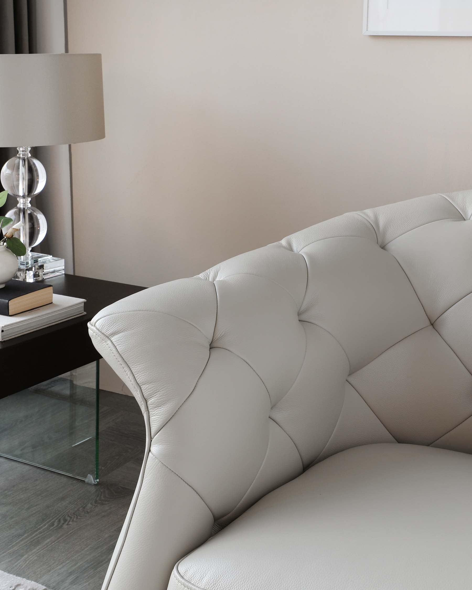 Elegant contemporary living room setting featuring a cream-colored tufted leather sofa with a plush back cushion. Beside it is a sleek black side table with a modern lamp, glass base, and neutral lampshade, complemented by a clear glass coffee table with a black frame in the foreground. The sophisticated and minimalist design creates a luxurious and comfortable ambiance.