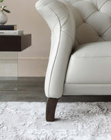 Close-up of an elegant cream-colored tufted leather sofa with dark wooden legs, partially visible beside a dark wooden side table on a textured white rug.