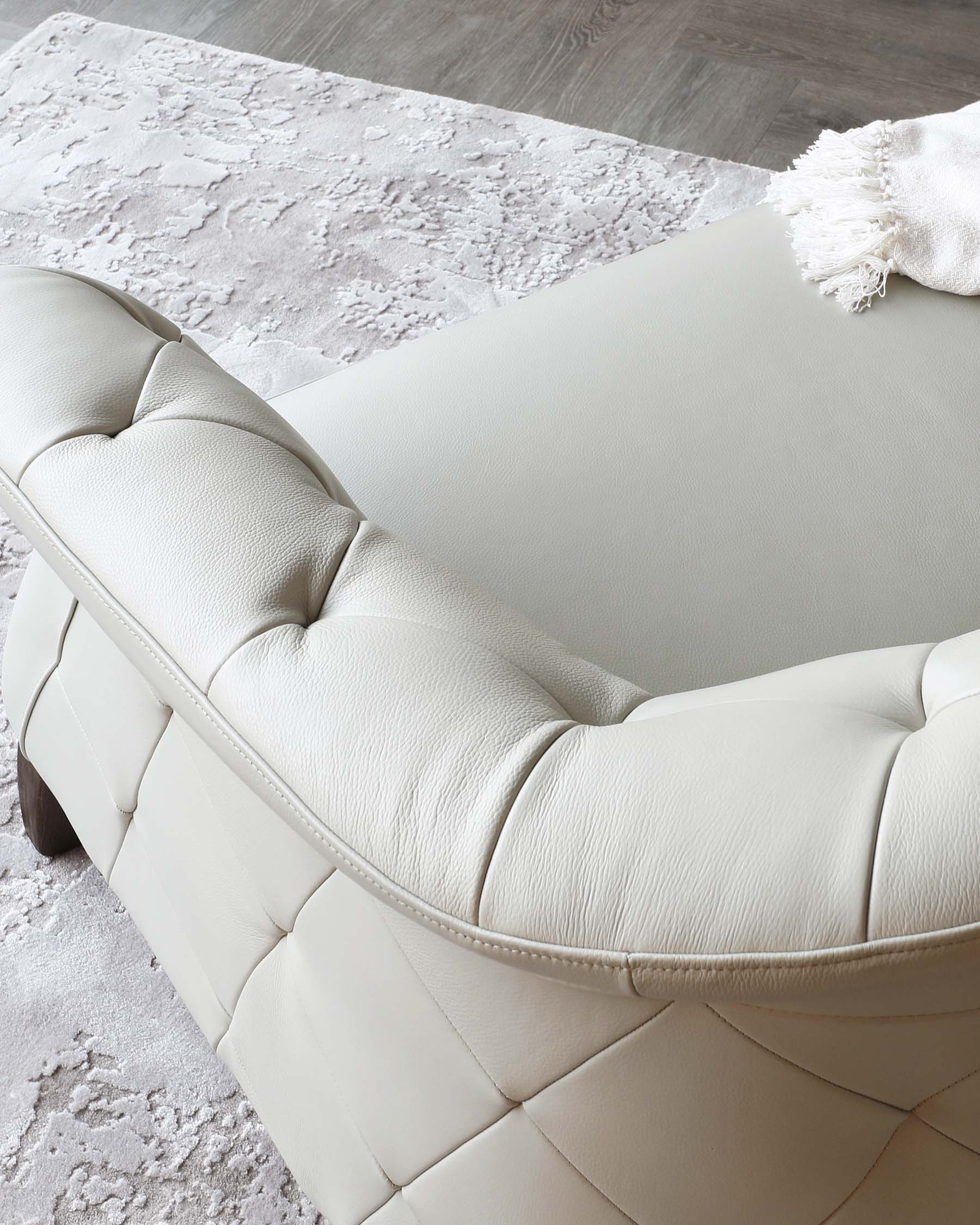 Close-up of a modern ivory leather sofa with a tufted back design and subtle stitching details, positioned on a textured light grey area rug over a hardwood floor. A corner of a white fringed throw blanket is delicately draped over one side of the sofa.