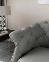 Elegant grey tufted sofa with a soft sheen, showcasing a classic curved silhouette and distinctive button tufting on the backrest. Beside the sofa is a clear glass side table with a modern chrome frame, supporting a chic table lamp with a translucent base and a clean white shade. A minimalist book and decorative vase with flowers add to the sophisticated ambiance.