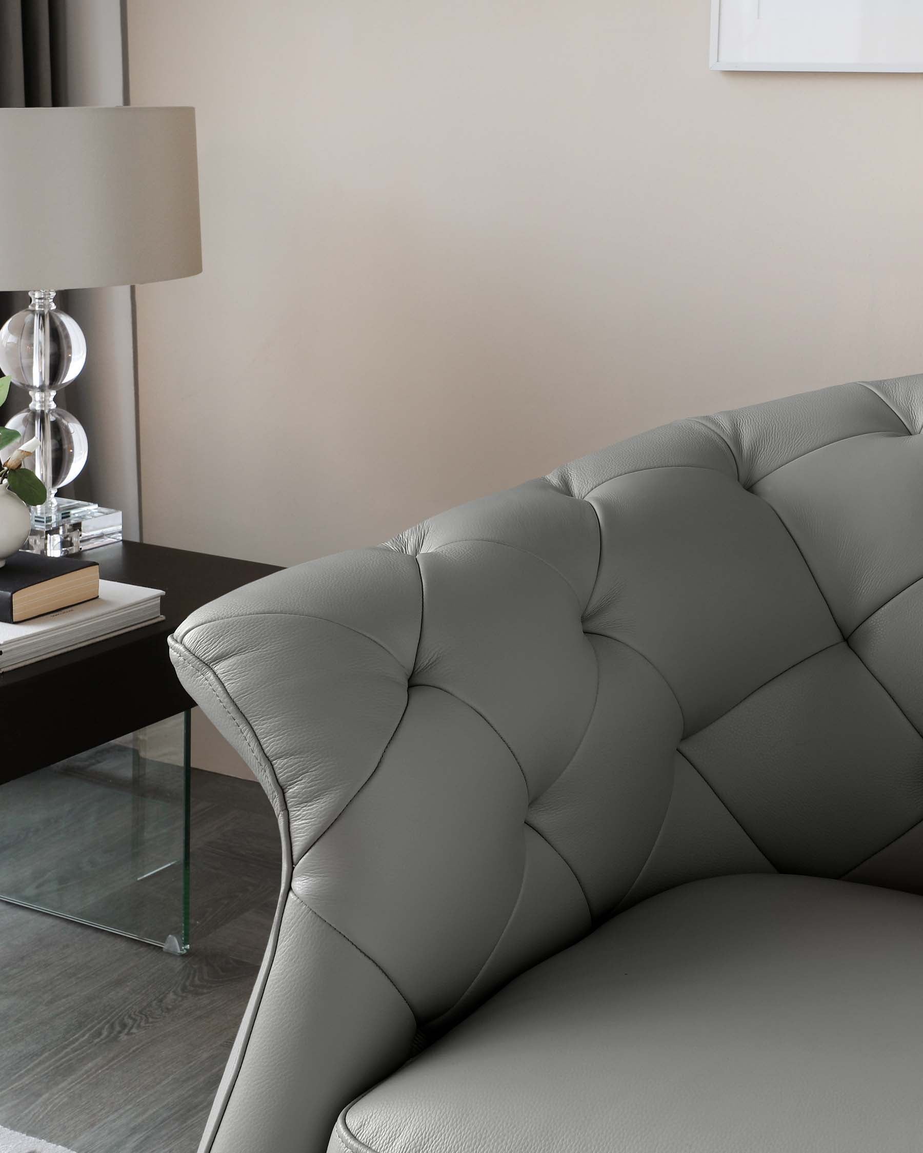 Elegant grey tufted sofa with a smooth, curving armrest and backrest design. Beside it, a minimalist clear glass side table with a lower shelf, hosting books and a small potted plant. A contemporary table lamp with a translucent base and a white shade provides ambient lighting, all set against a neutral wall backdrop.