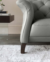 Elegant light grey tufted leather sofa with a classic design featuring a rounded armrest, high back, and dark wooden legs, partially shown against a neutral backdrop, with a dark wooden side table displaying books and a potted plant, all atop a textured white area rug.