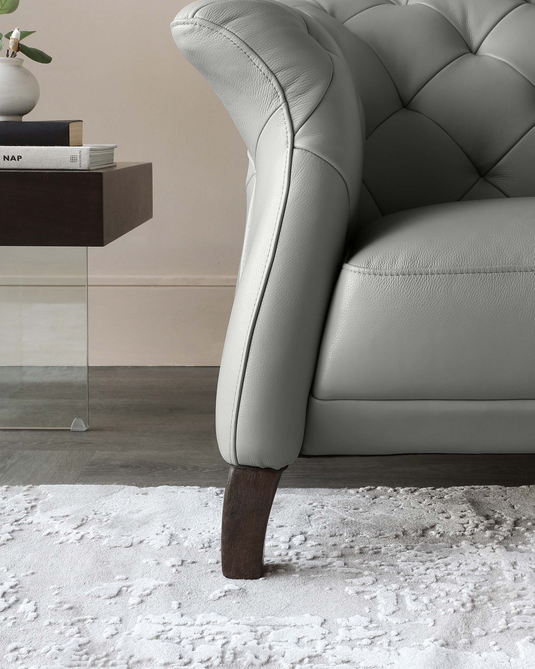 Elegant light grey tufted leather sofa with a high back and rounded arms featuring dark wooden tapered legs, accompanied by a modern dark wood side table with a simple vase on it, over a textured white area rug.