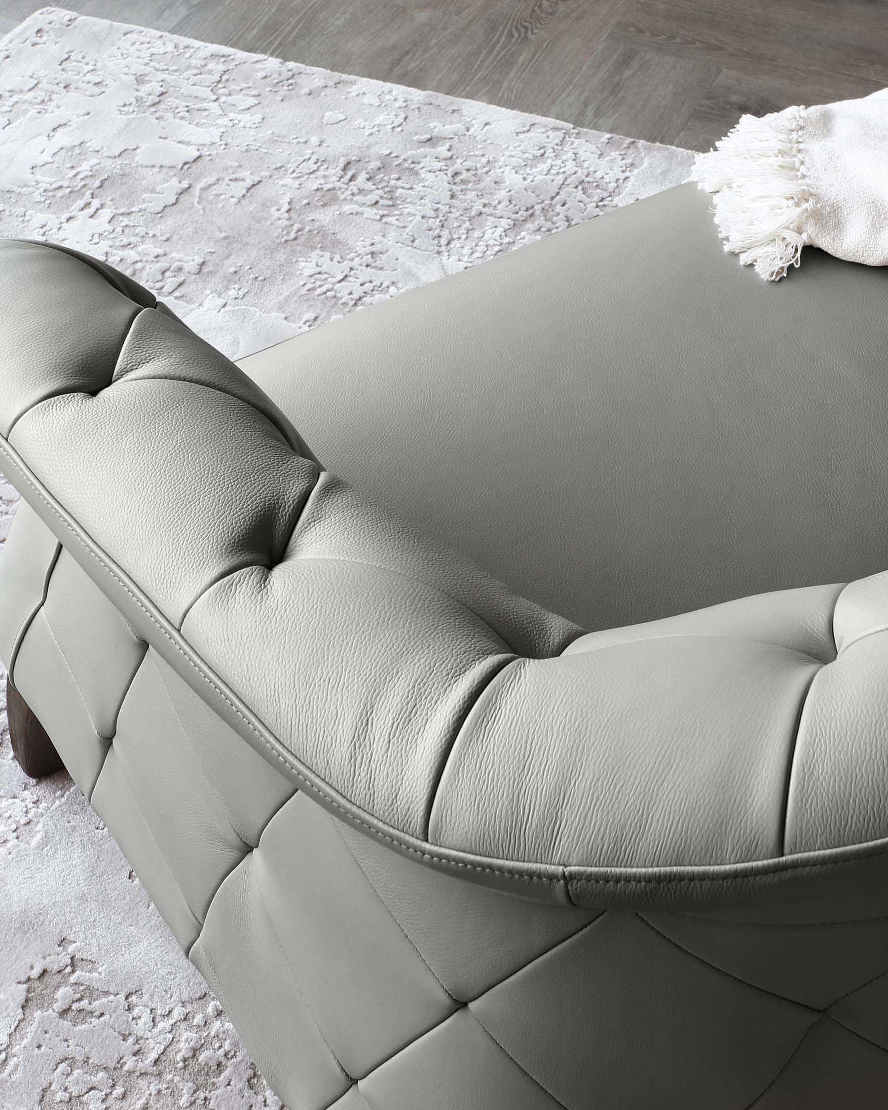 Elegant modern grey leather sofa with tufted upholstery and clean, stitched lines, resting on dark wooden legs, accompanied by a soft off-white textured throw blanket draped over one corner. Positioned on a plush white and grey area rug with abstract pattern in a room with hardwood flooring.