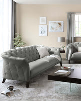 Elegant living room setup featuring a plush, tufted-back grey sofa with curved armrests and a matching armchair. A dark wooden rectangular coffee table with a clear glass centrepiece sits atop a textured white area rug. Beside the sofa is a dark side table with a sleek, silver lamp.