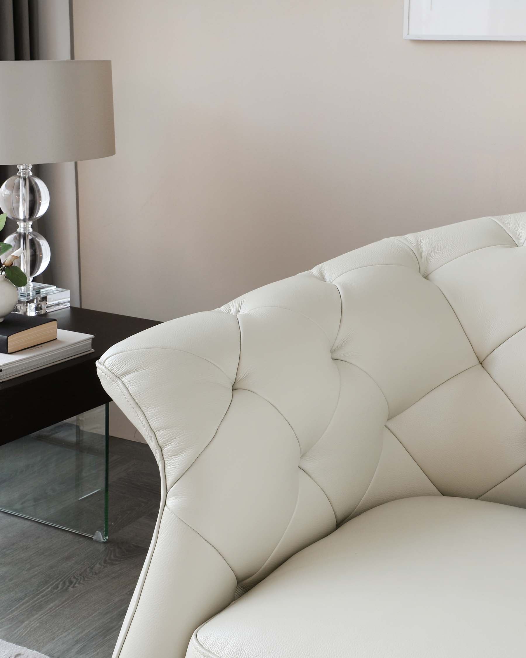 Elegant cream tufted leather sofa with a modern glass side table featuring a transparent base and black surface, accompanied by a stylish lamp with a clear base and white shade.