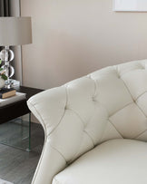 Elegant cream tufted leather sofa with a modern side table featuring a clear glass top, black frame, decorative table lamp, and a neatly stacked collection of books.