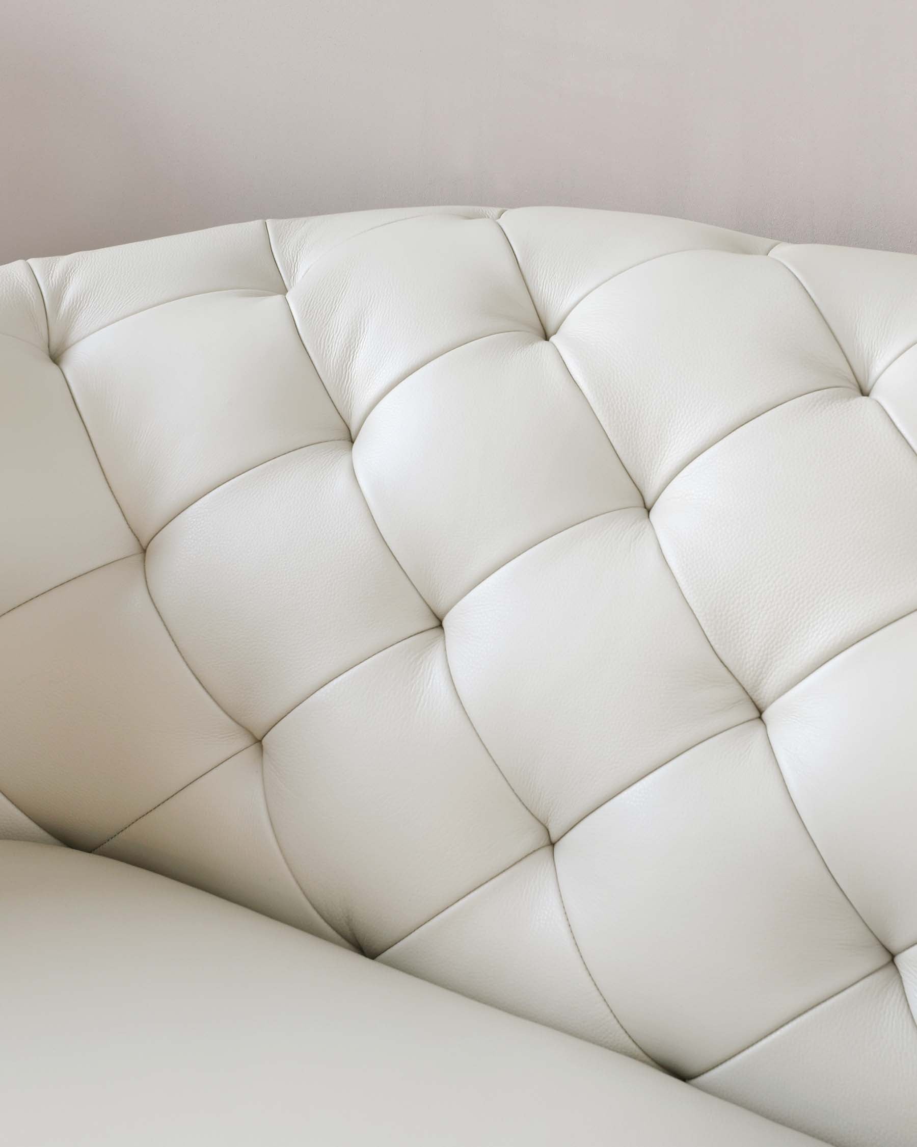 Close-up of an ivory leather tufted sofa with a plush design and elegant stitching detail, against a soft grey background.