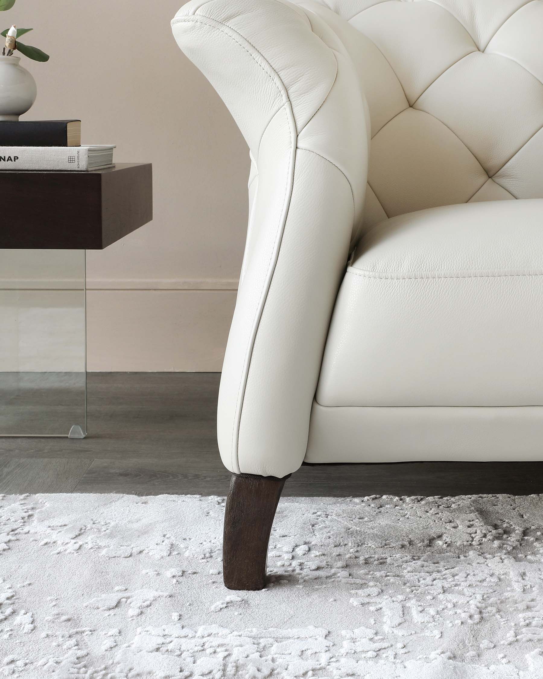 Close-up of a creamy white tufted leather sofa showing a detailed view of the plush upholstery and dark wooden leg, placed on a textured white rug with a minimalist dark wooden side table holding books and a small plant in the background.