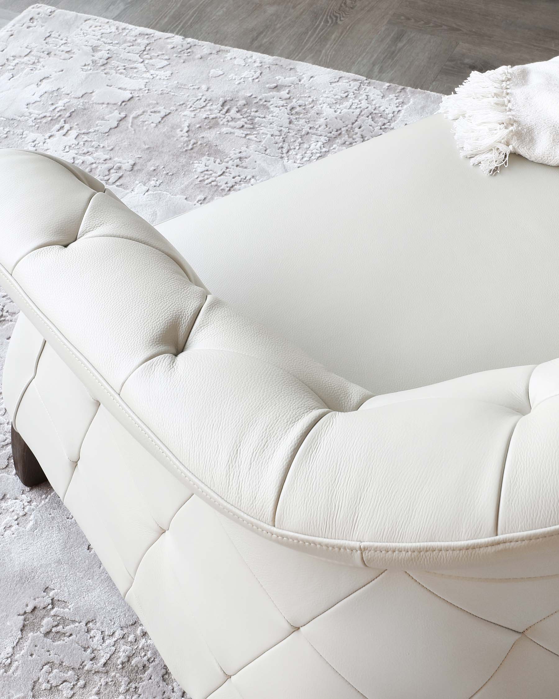 Elegant modern leather sofa in a pristine white colour with a tufted backrest and seat design, dark wooden legs partially visible. A cosy off-white throw blanket with tassels is draped on one side of the sofa. The sofa is positioned on a plush grey area rug with a textured pattern, suggesting a luxurious and contemporary living space.