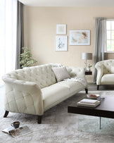 Elegant cream-colored tufted sofa with matching armchair featuring classic rolled arms and deep button tufting detail, accompanied by a modern rectangular glass coffee table with a dark wooden base. A distressed white patterned area rug rests beneath the furniture set.