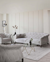 Elegant living room furniture set featuring a curved light grey tufted sofa with velvet upholstery, complemented by two matching armchairs with a diamond tufting design. A round glass-top coffee table with a unique golden geometric base sits on a soft white area rug, anchoring the arrangement. The overall aesthetic is chic and sophisticated with a contemporary twist.