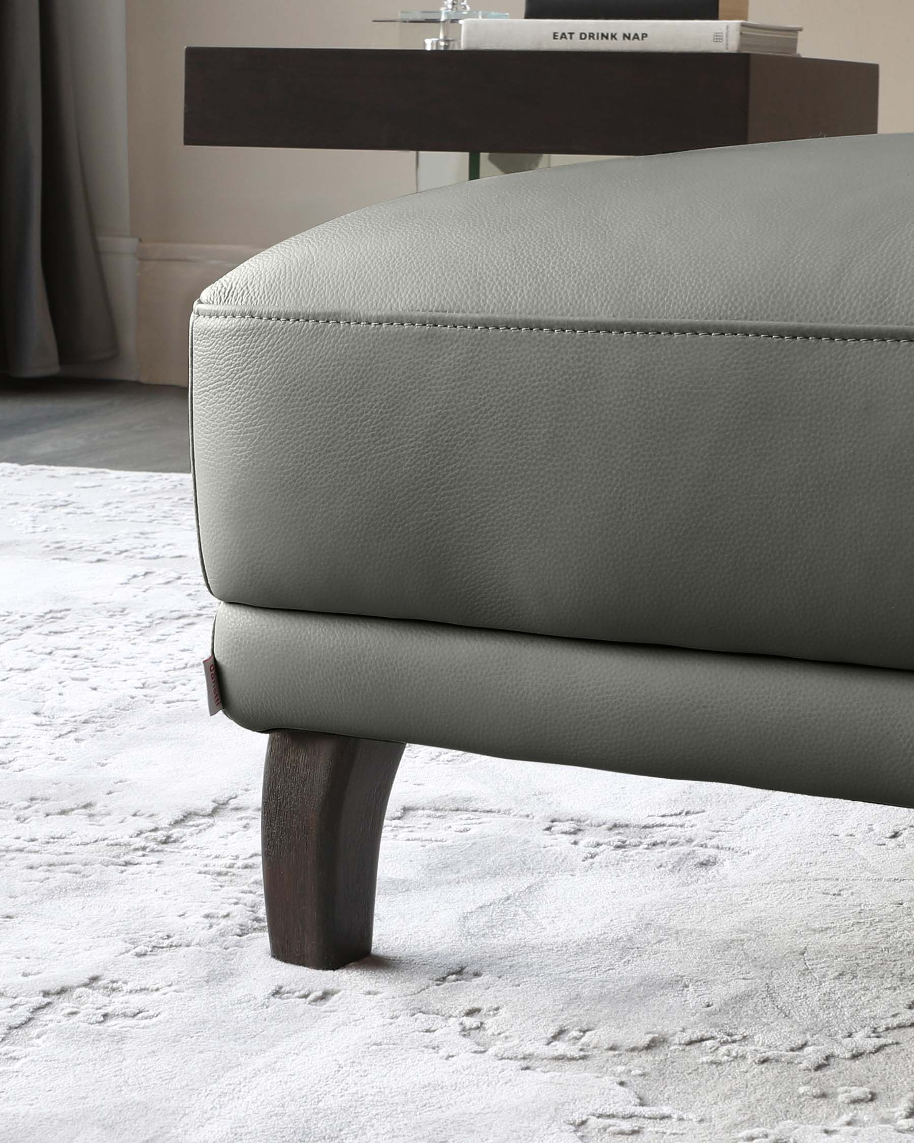 Close-up of a modern light grey leather ottoman with a delicate stitching detail, resting on dark wooden tapered legs. In the background, there is a sleek dark wood sideboard with a book titled "EAT DRINK NAP" on top, set against a neutral toned wall with minimalist decor.