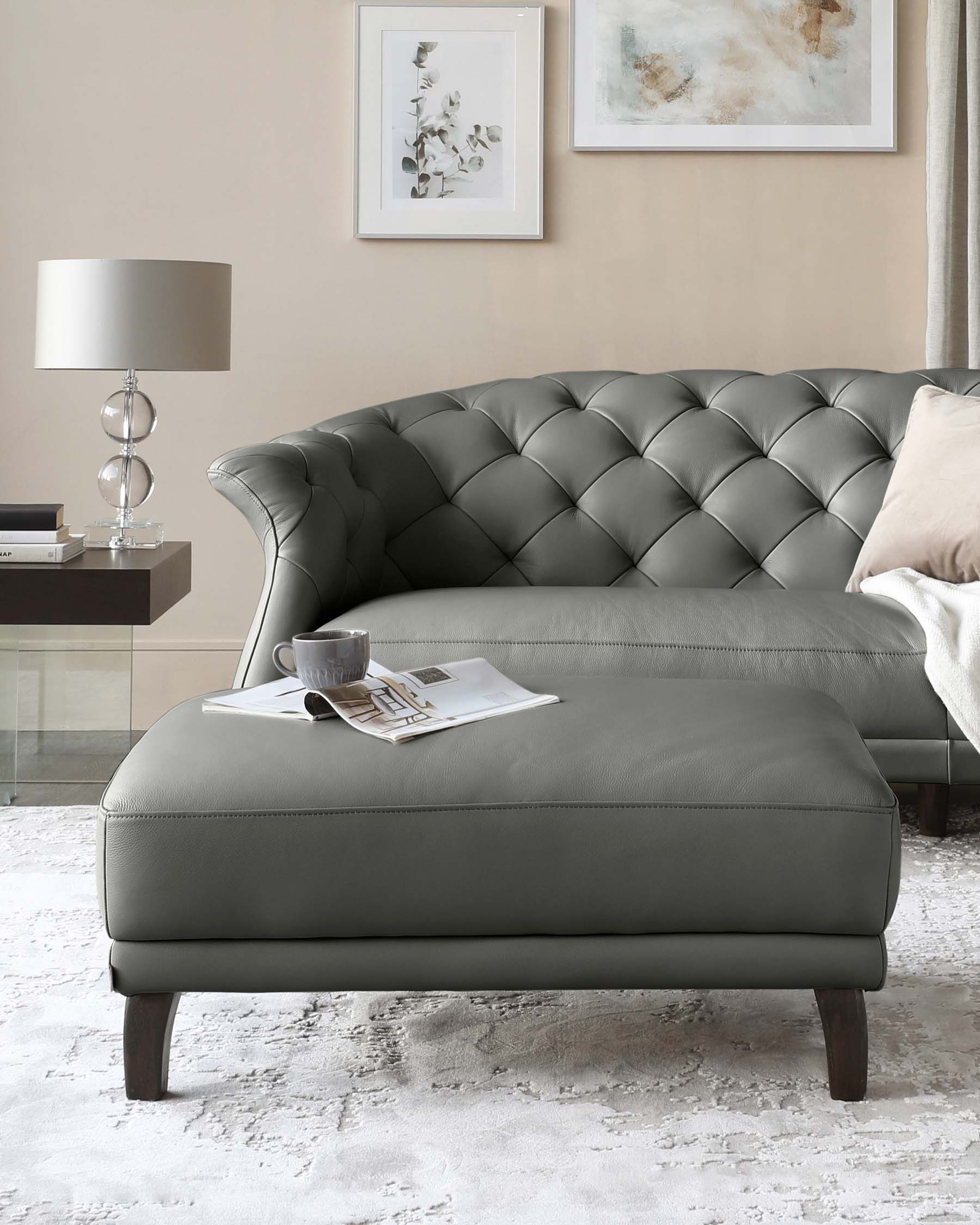 Elegant dark grey tufted chaise longue with matching ottoman, featuring button detailing and dark wooden legs, complemented by neutral-toned pillows, and set against a soft cream wall with tasteful art and accent lighting. On the ottoman, a cup and saucer set alongside a magazine creates a cosy reading nook atmosphere.