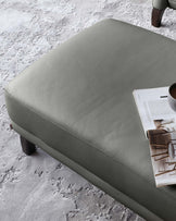 Modern grey leather ottoman with visible stitching detail and tapered wooden legs, placed on a textured grey area rug, with an open magazine on top.