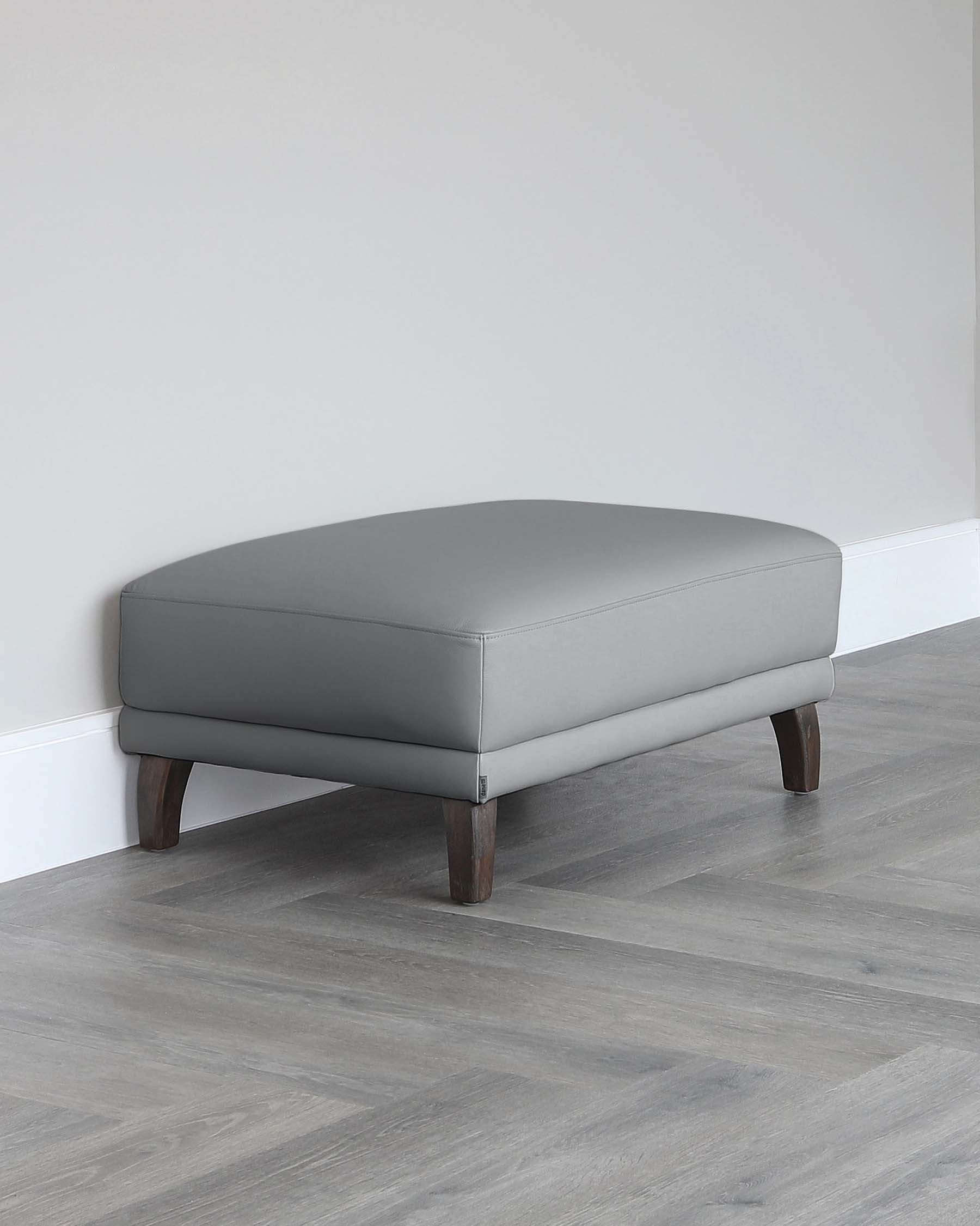 Modern grey upholstered ottoman with a sleek design and dark wooden legs, set against a muted grey wooden floor and a plain light-coloured wall.