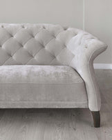 Elegant grey fabric sofa with tufted backrest and scrolling arms, featuring dark-stained wooden legs, placed on a light wooden floor against a grey wall.