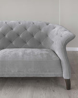 An elegant grey fabric loveseat with tufted backrest and curved armrests, standing on dark wooden legs against a light wall on a wooden floor.