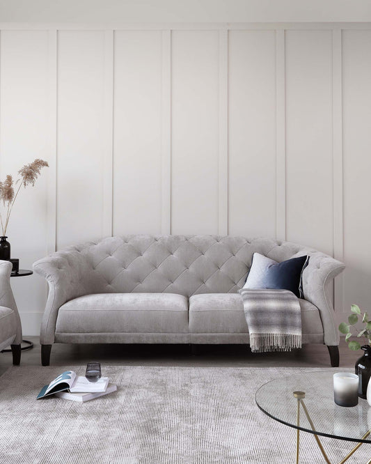 Elegant light grey tufted fabric sofa with rolled arms and a curved, button-tufted backrest. Two round side tables in black, one with a vase of pampas grass, the other with books and a glass. A grey woven area rug underfoot and a round coffee table with a gold frame and glass top complete the scene.