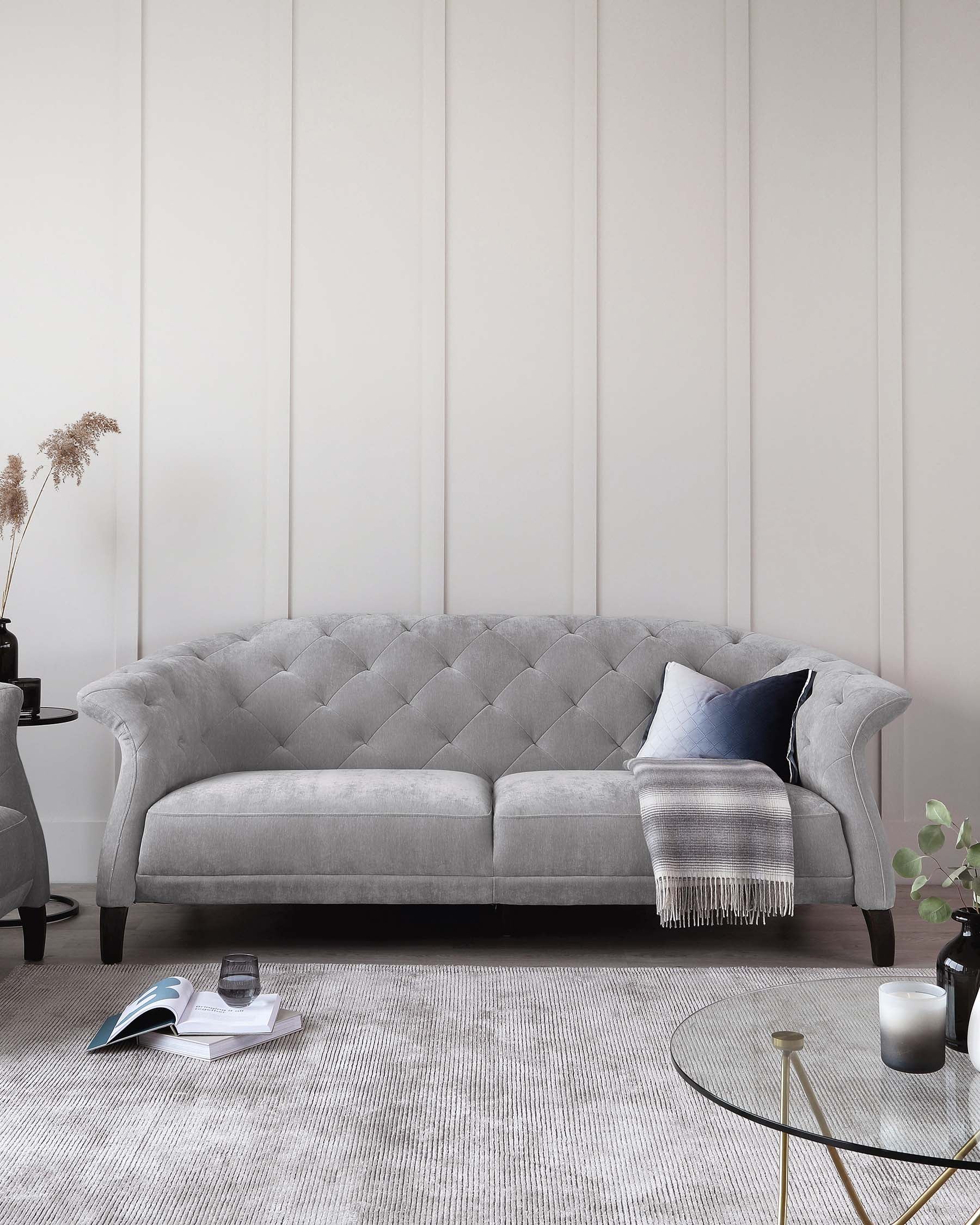 Elegant grey tufted chesterfield sofa with plush upholstery, accompanied by a round gold and glass coffee table, resting on a textured off-white area rug. Decorative accents include a dark side table with vase, books, candle, and a cosy throw blanket draped over the sofa.