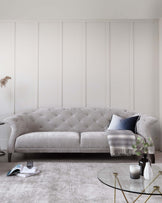 Elegant light grey tufted fabric sofa with a curved backrest, plush seating, and fine detailing on a textured light-coloured rug, complemented by a round glass-top coffee table with a slender golden frame. The setting is accented by small decorative pieces, including vases with greenery, a throw blanket, and a cushion.