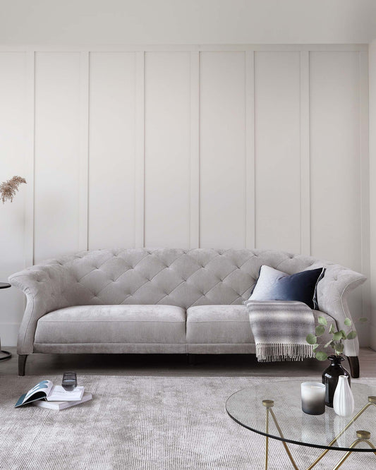 Elegant light grey tufted fabric sofa with a curved backrest, plush seating, and fine detailing on a textured light-coloured rug, complemented by a round glass-top coffee table with a slender golden frame. The setting is accented by small decorative pieces, including vases with greenery, a throw blanket, and a cushion.