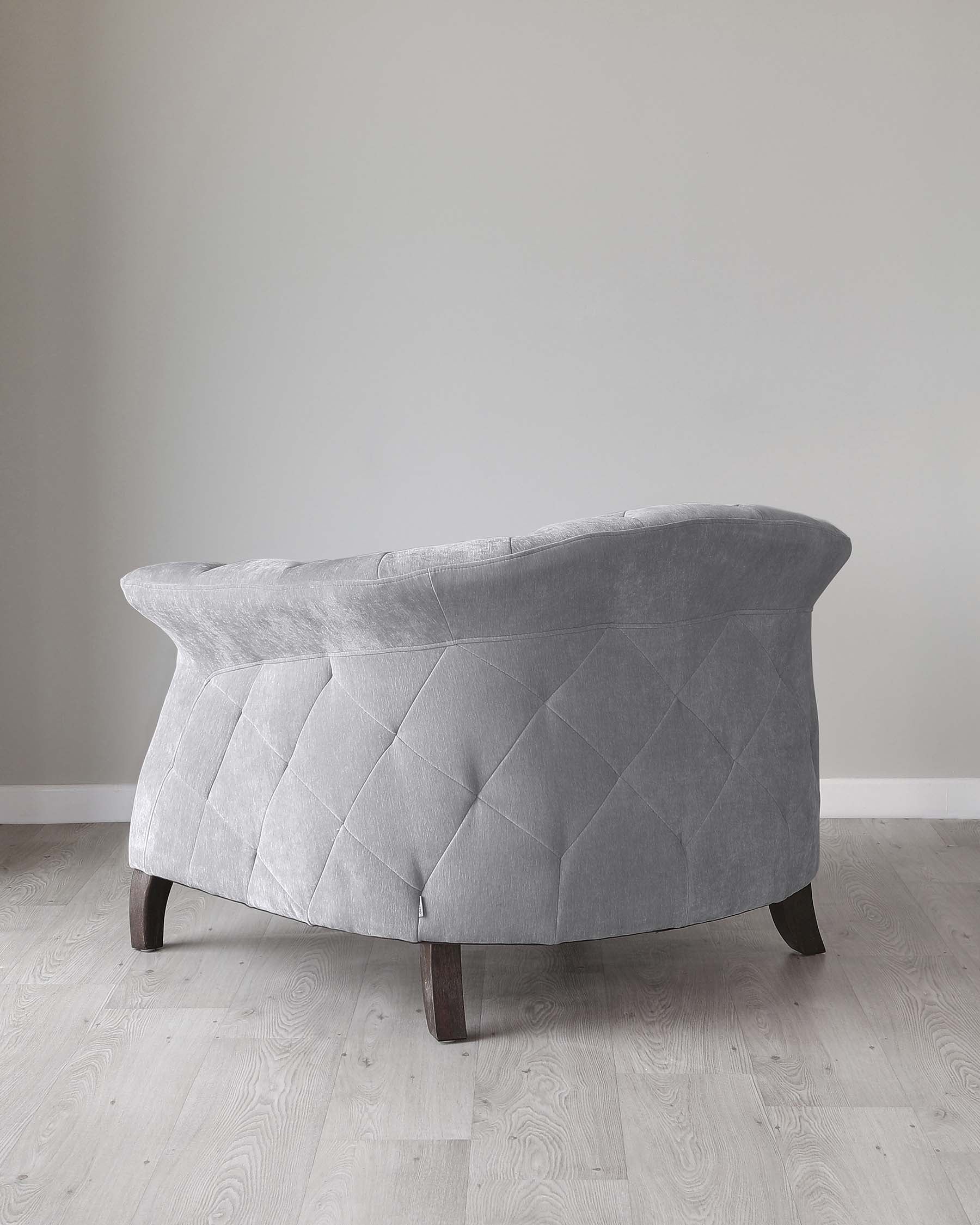 Elegant grey fabric loveseat with a tufted, quilt-patterned backrest and seat, featuring dark wooden legs, set against a neutral wall on a light wooden floor.