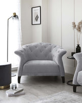 Elegant grey tufted loveseat with curved backrest and armrests, featuring dark wooden legs. The room is accessorized with a round marble-top side table with a gold and black base, a floor lamp with a minimalist design, and a decorative vase with dried pampas grass. A framed abstract art piece hangs on the wall, enhancing the contemporary chic aesthetic of the space.