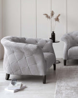 Elegant light grey tufted armchair with a plush, diamond-pattern design. The chair features a wide, comfortable seat and a curved backrest, supported by dark wooden legs, complementing a matching sofa in a neutral-toned room with a textured grey rug, a small black side table, and decorative pampas grass in a slender vase.