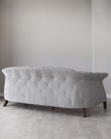 Elegant curved sofa with tufted light grey upholstery and dark wooden legs, featuring a distinctive diamond pattern on its back and seat. Positioned in a minimalist room with light wooden flooring and white walls.