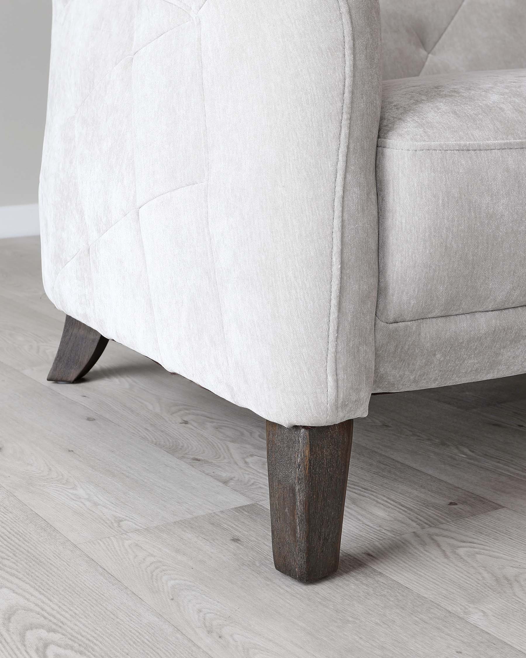 Close-up of a contemporary style armchair with light beige upholstery and tufted backrest detailing. The chair stands on angled, dark-stained wooden legs that contrast with the light-coloured fabric, and the flooring is a light grey wood laminate.