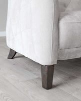 Close-up view of a light beige fabric upholstered armchair with a tufted design on the side panel and sturdy, angled, dark wooden legs set on a light hardwood floor.