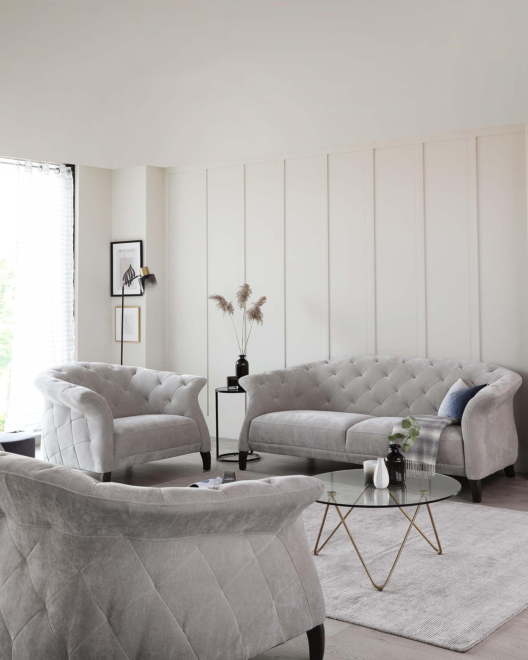 Elegant living room furniture set featuring a tufted grey velvet sofa with a curved back, matching tufted armchairs with a classical design, a circular glass-top coffee table with a golden frame, and a rectangular side table with a marble top and a black frame. A soft white area rug under the coffee table adds warmth to the setup.