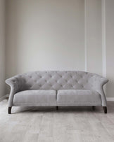 Elegant grey fabric sofa with tufted backrest and gently curved armrests, featuring dark wooden legs, set against a neutral wall and light wood flooring.