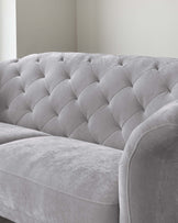 Elegant grey upholstered sofa featuring a tufted backrest with a diamond pattern design, plush seat cushions, and a smooth, curved armrest on one side. The sofa is placed against a neutral-coloured wall, highlighting its chic and sophisticated style.